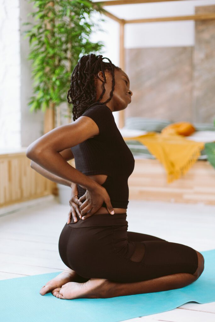 Vertical African American multicultural sportswoman with dreadlocks stretching, inclining, do yoga. Female holding back, feeling pain in joints and muscles, backache. Health and body care. Side view