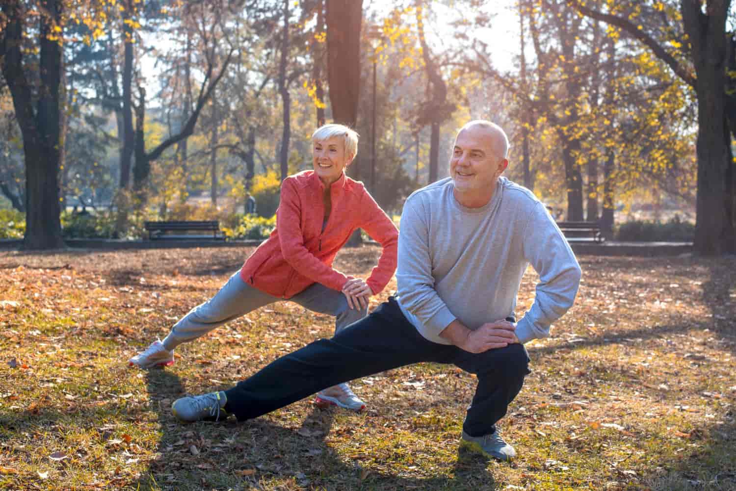 Senior couple stretching outdoors in a park, performing side lunges and enjoying their fitness routine in the fresh air.