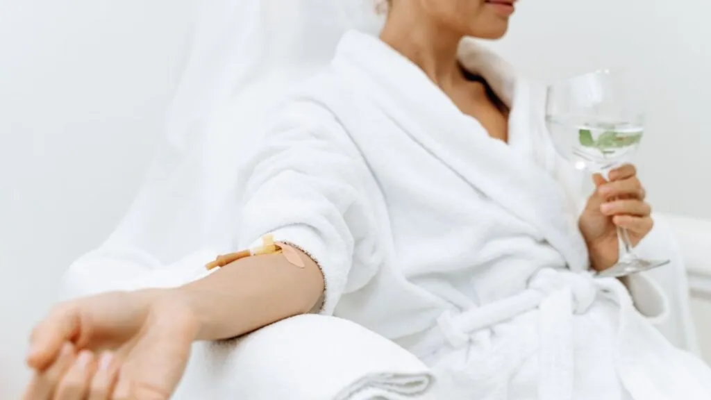 Person in a white robe receiving IV therapy while holding a glass of water